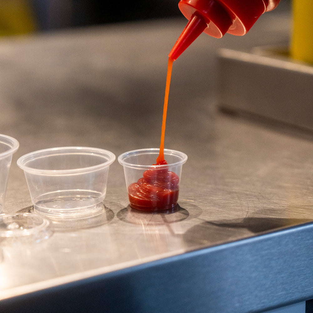 Packaging Wise 1oz clear PET sauce pot — ideal portion cup for ketchup, dips & condiments, shown being filled at counter service.